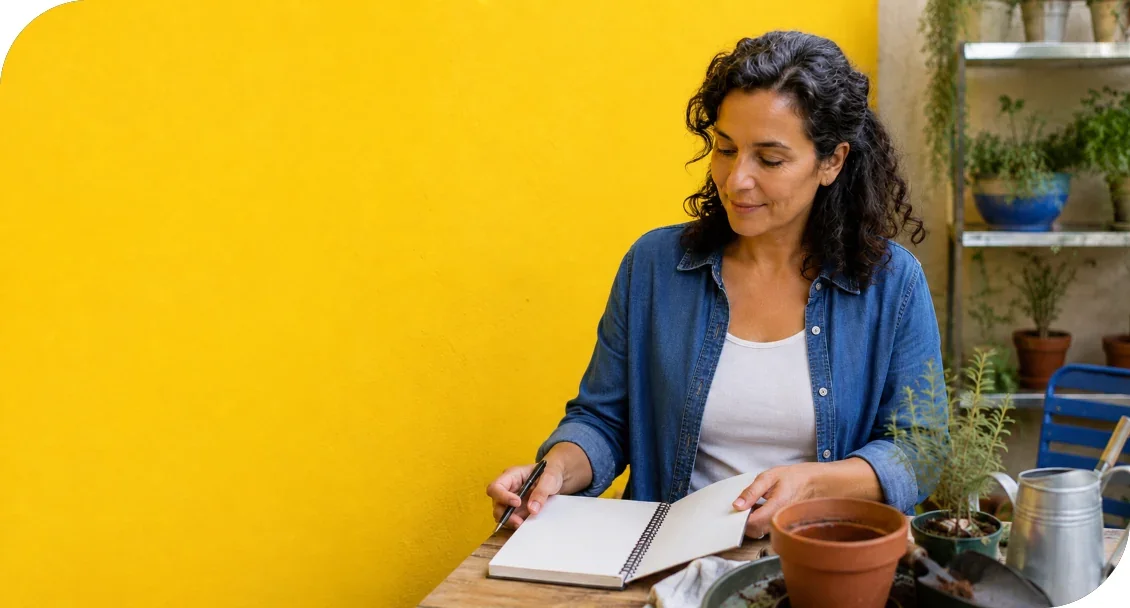 A woman writing in a notebook beside gardening supplies and potted plants.