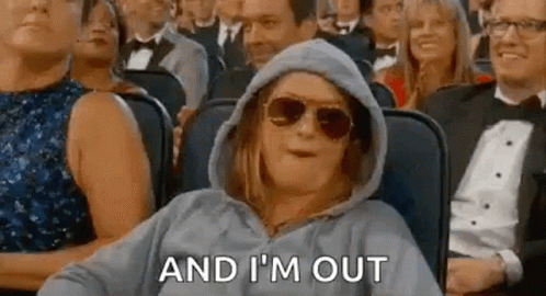 A woman in a sweatshirt and sunglasses sitting in the audience saying "I'm Out" 
