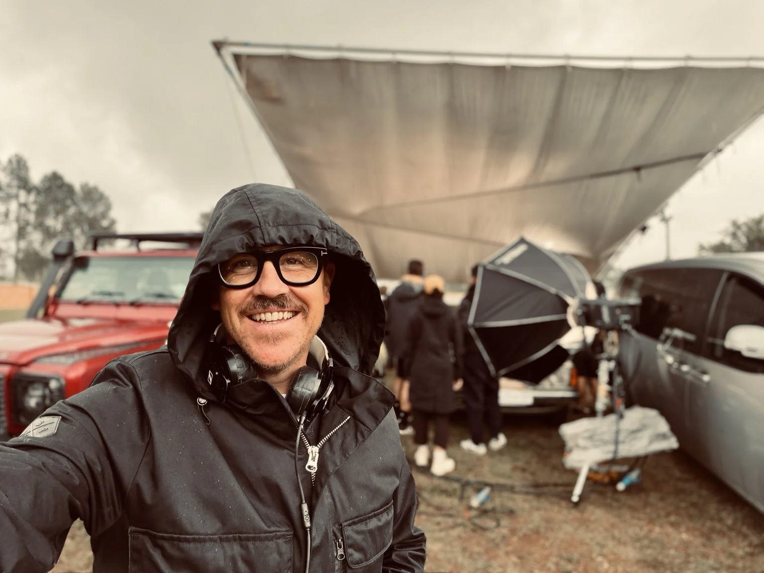 Jono Hall on set, smiling beneath a rain hood