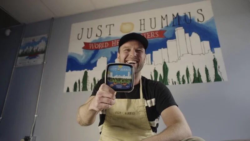 Colin, Founder of Just Us Foods sits in the original production facility while smiling and holding up a tub of original Just Hummus