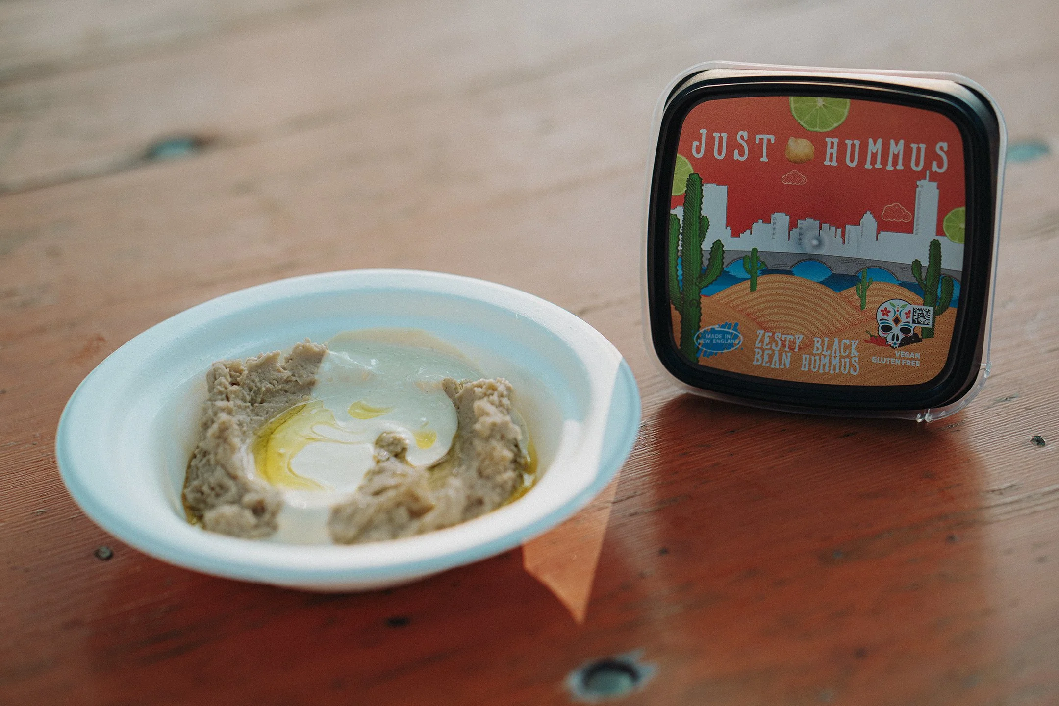 Zesty Black Bean Just Hummus packaging shown next to a bowl of Just Hummus topped with tahini dressing and olive oil