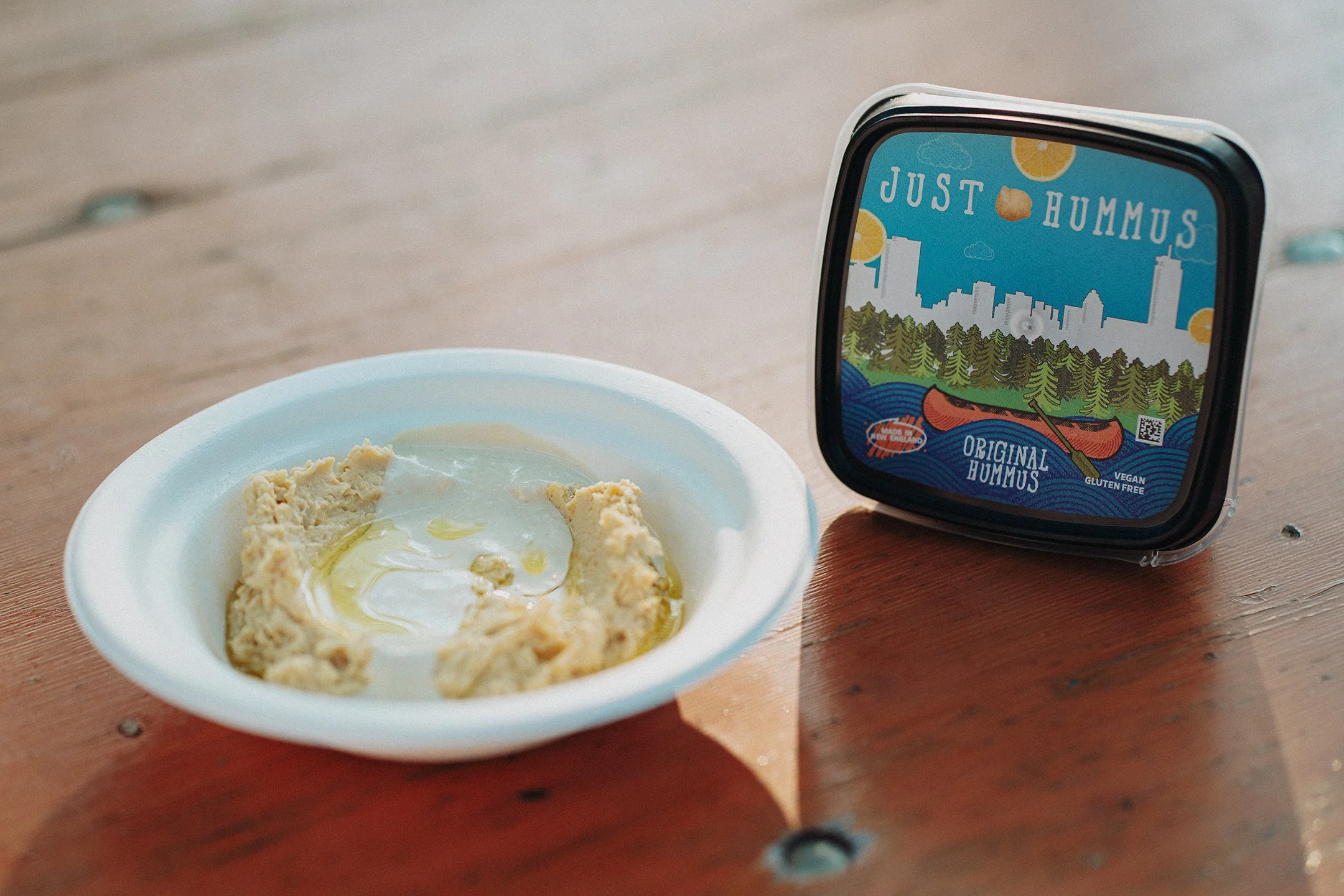 Original Just Hummus packaging shown next to a bowl of Just Hummus topped with tahini dressing and olive oil