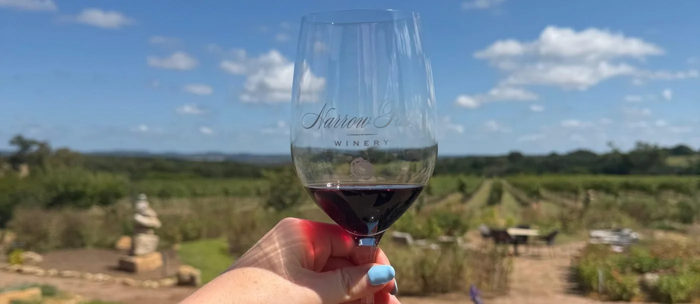 A glass of Texas red held up over the vine rows and lavender at Narrow Path Winery near Stonewall