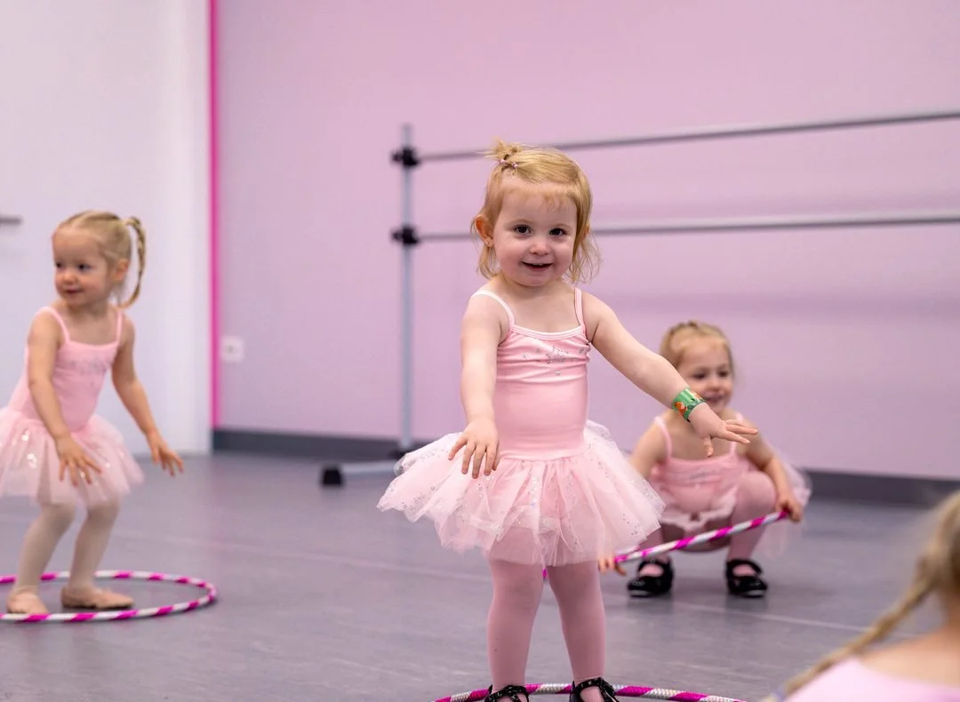 Preschool dancers in pink tutus balancing inside hula hoops during a creative movement class