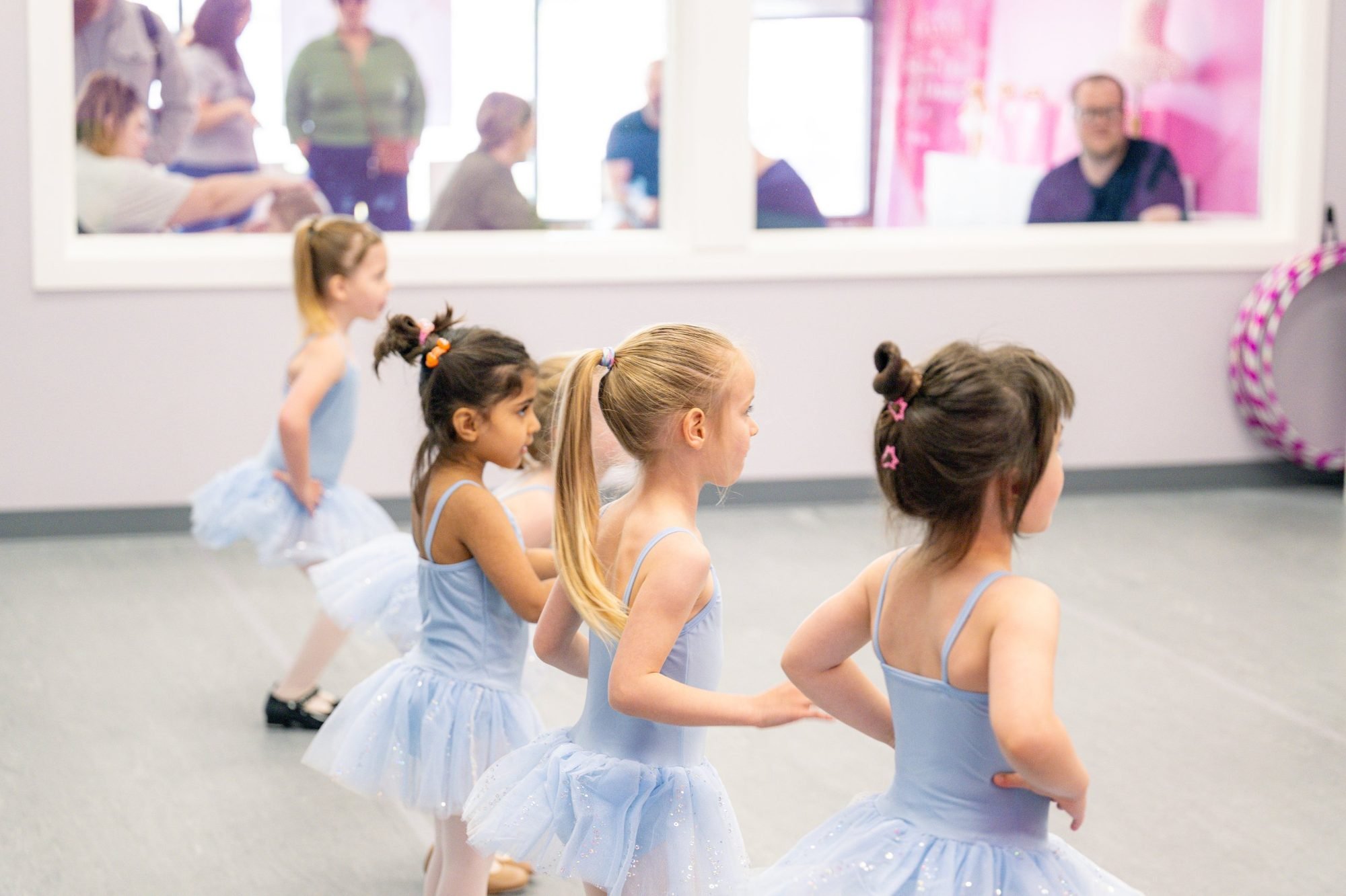 Young dancers in blue leotards and tutus during class, with parents watching through the lobby viewing windows