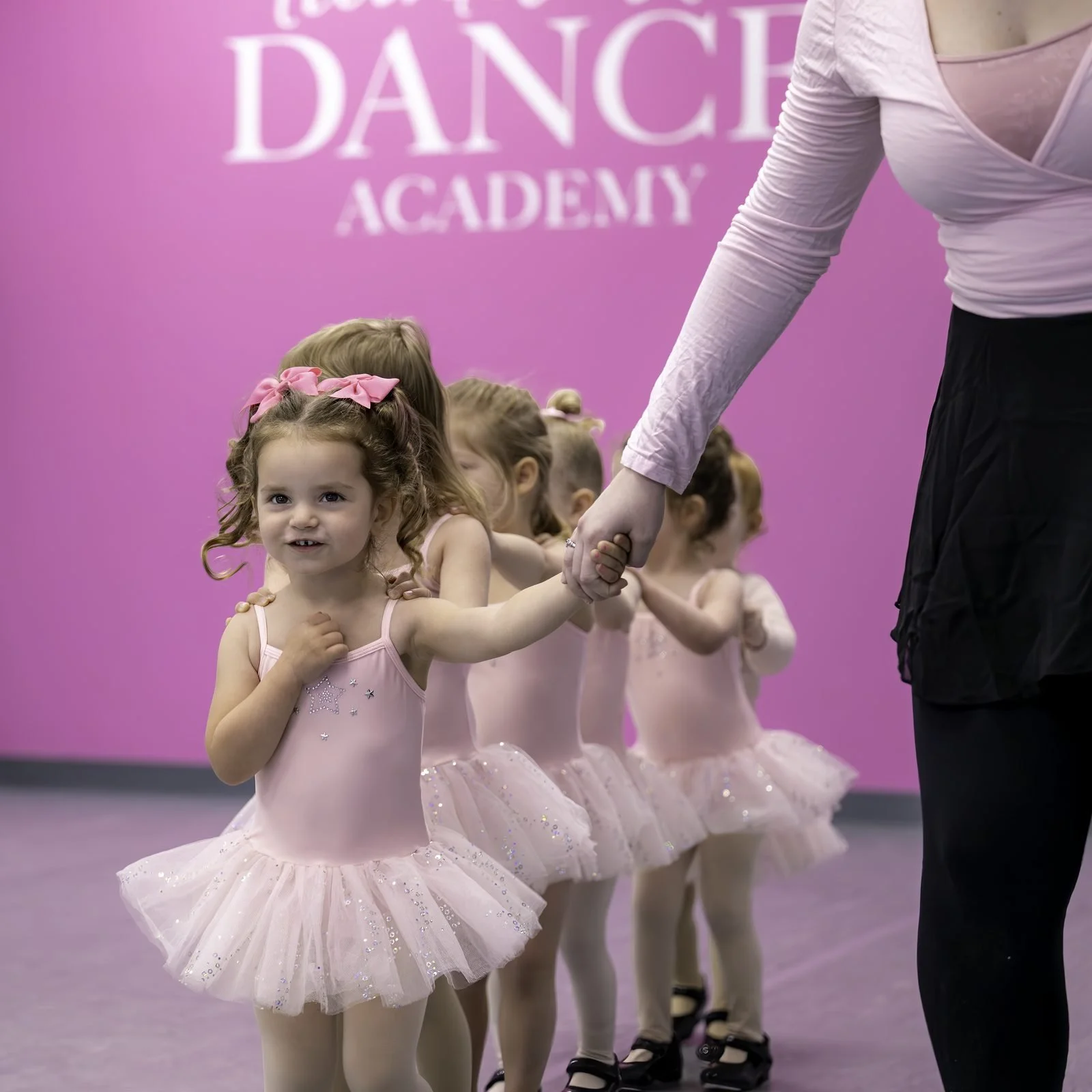 A teacher leads a line of toddlers in pink tutus across the studio