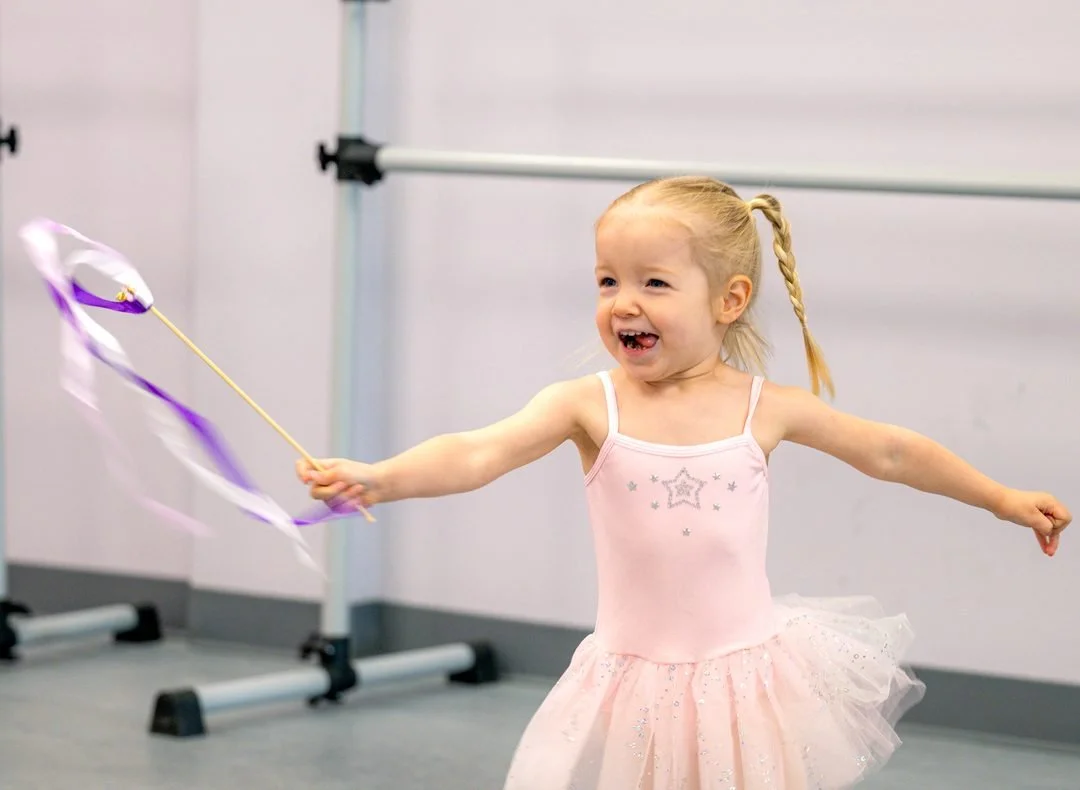 Smiling preschool dancer in a pink tutu twirling a purple ribbon wand