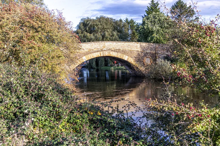 An old stone bridge over the upper River Thames in the Oxfordshire countryside, framed by autumn hedgerows