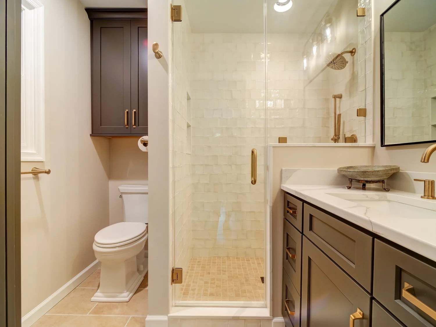 Bathroom with a glass-door shower in cream handmade-look tile, brass fixtures, a dark vanity with a marble top, and a separate compartmented toilet area behind a doorway