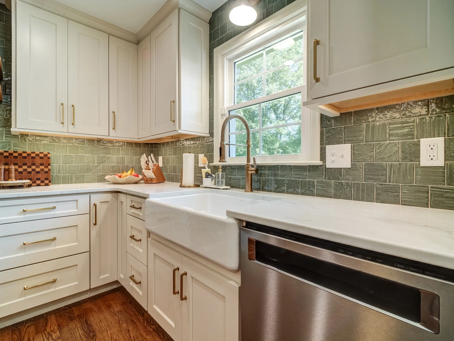 White shaker cabinets with a green handmade-look tile backsplash, white farmhouse apron sink, brass gooseneck faucet, and marble-look quartz counters