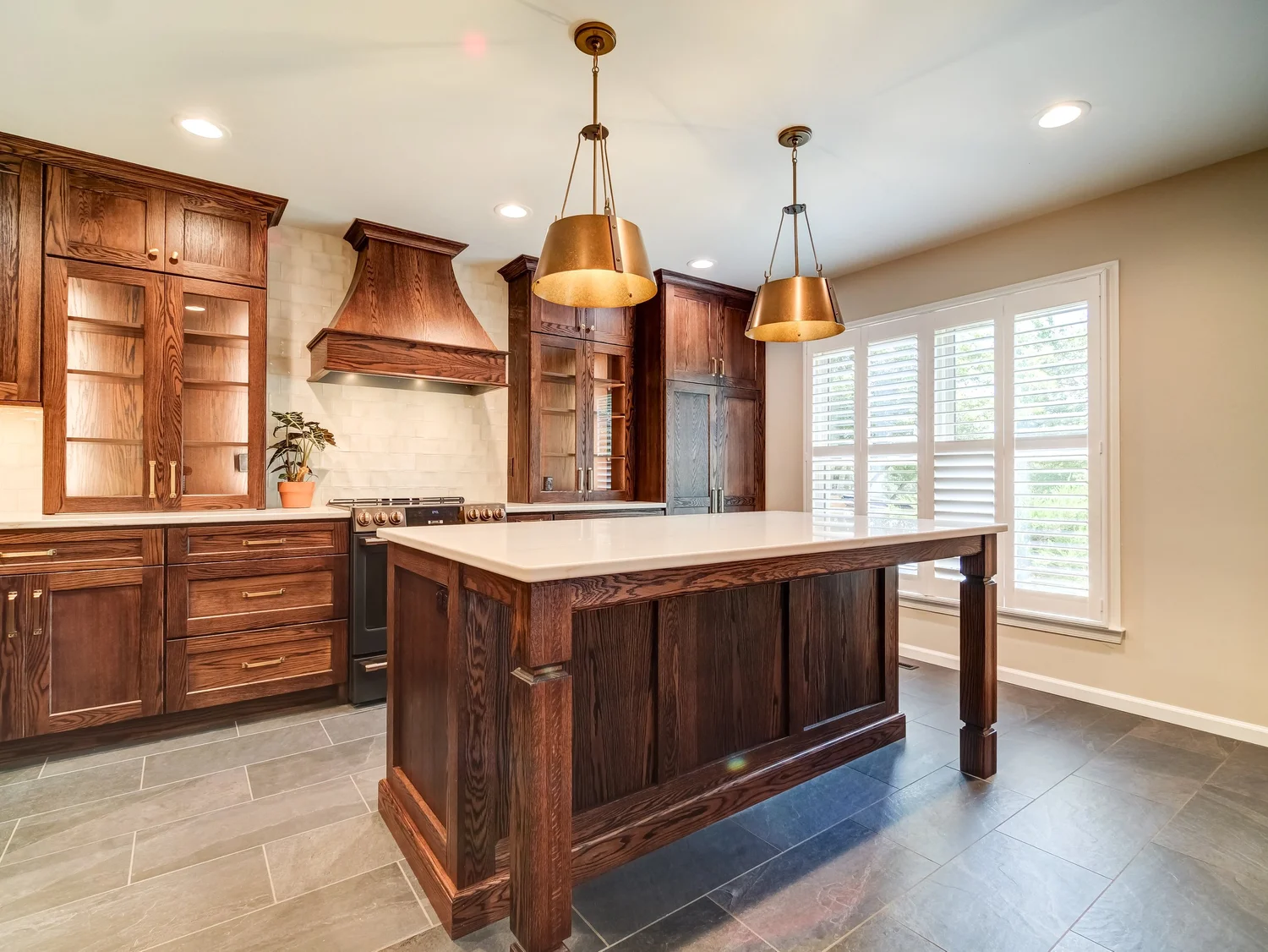 Dark stained oak kitchen cabinetry with a custom wood range hood, glass-front uppers, and a large quartz-topped island lit by two brass pendants