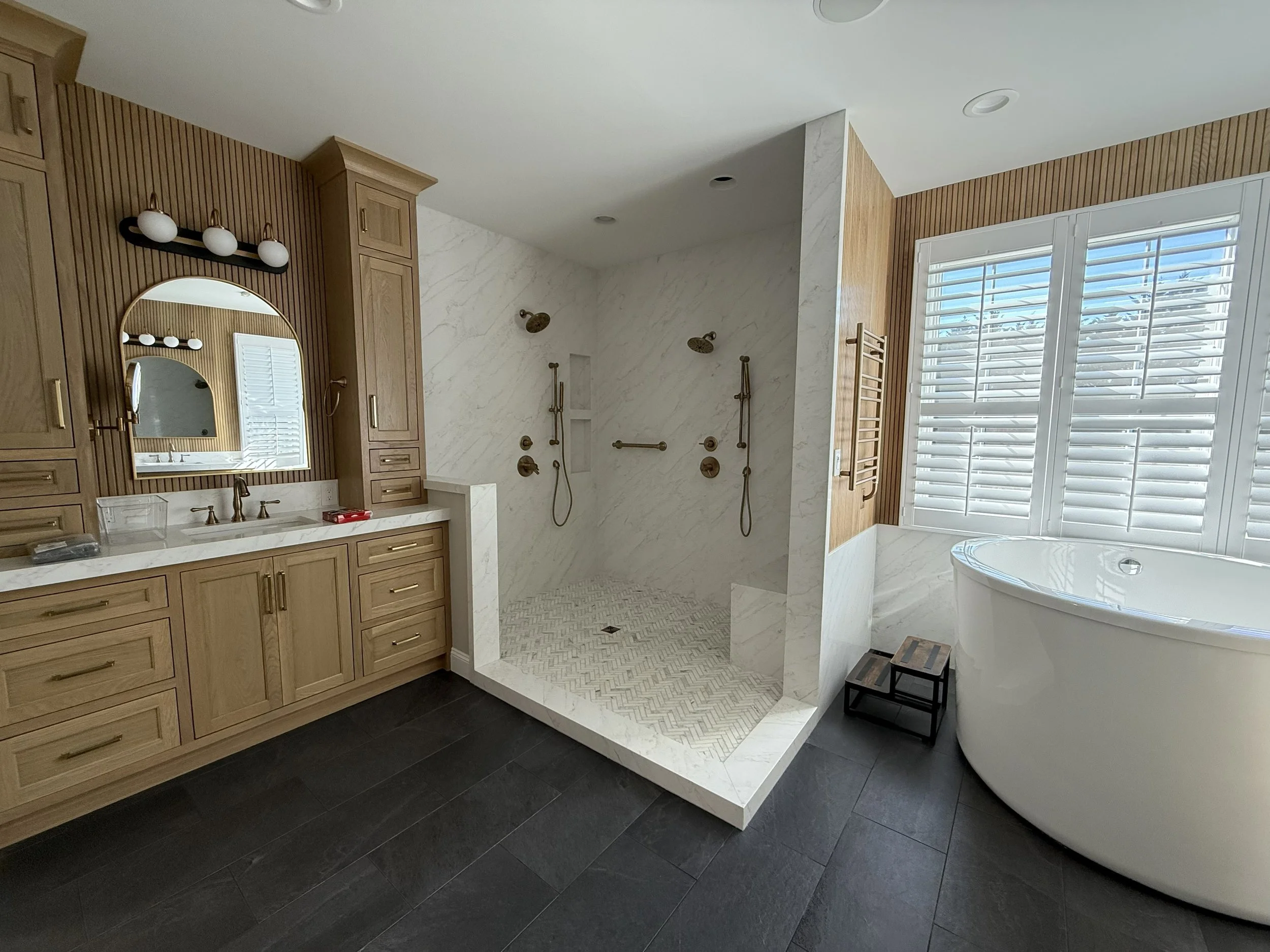 Primary bathroom with white oak fluted paneling, a floor-to-ceiling marble slab shower with two shower heads and a herringbone floor, a freestanding tub, brass fixtures and a towel warmer