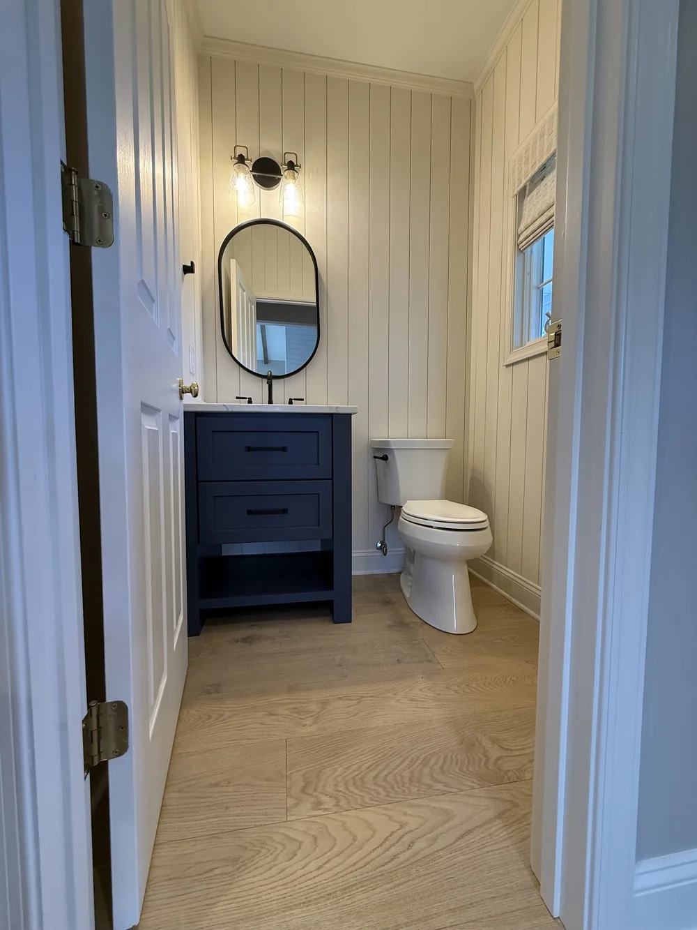 Small powder room with a navy vanity, cream vertical shiplap paneling, an oval black-framed mirror and wide-plank wood-look flooring