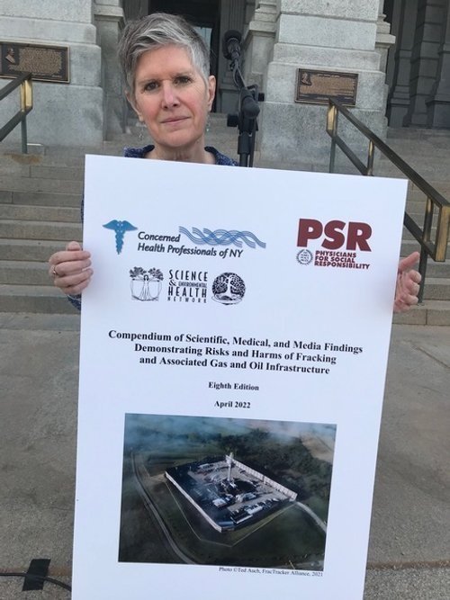 Dr. Sandra Steingraber holds a large, white poster with logos at the top for 3 different organizations with the title "Compendium of Scientific, Medical, and Media Findings Demonstrating Risks and Harms of Fracking" and a picture of a fracking site. 