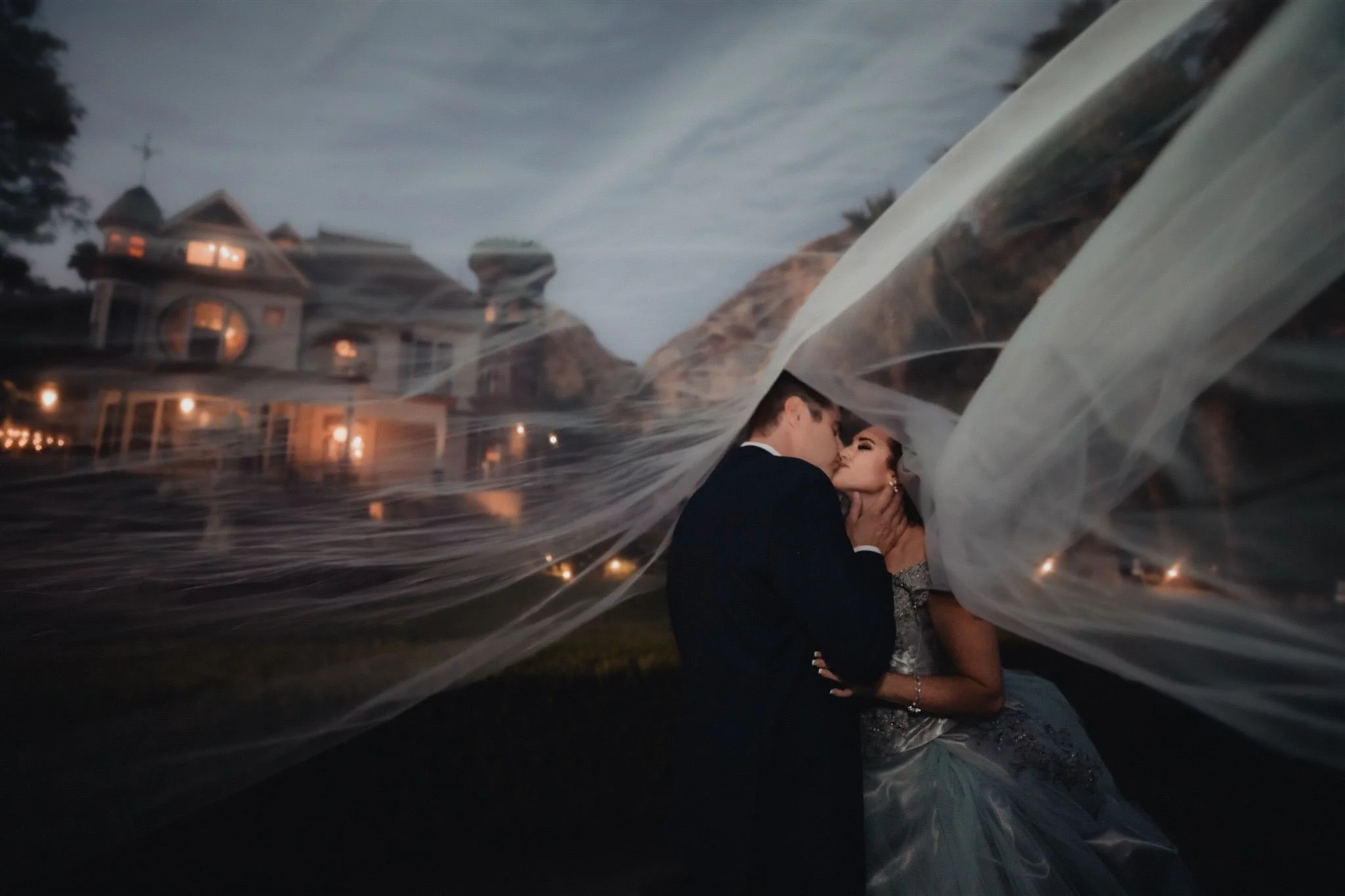 Bride and groom sharing a kiss under a flowing veil with Newhall Mansion glowing behind them at twilight