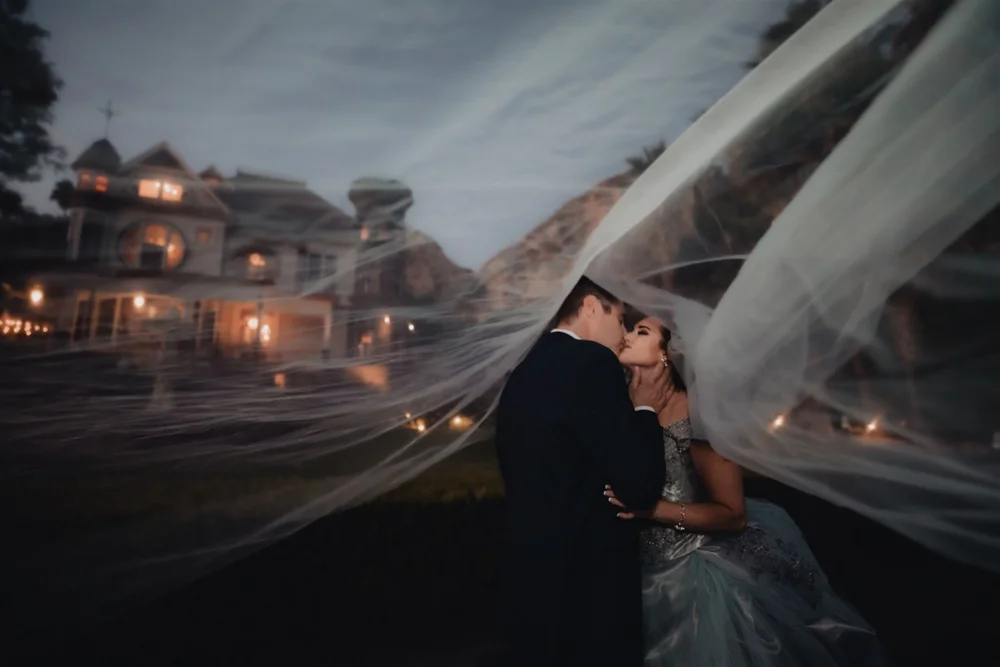 Bride and groom sharing a kiss under a flowing veil with Newhall Mansion glowing behind them at twilight