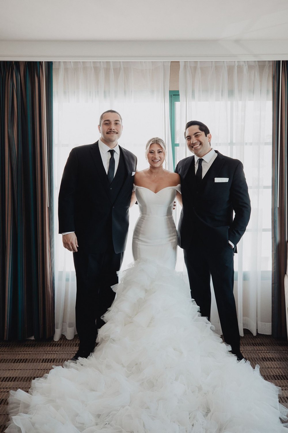 Bride in a heavily ruffled mermaid gown with a cathedral train standing between two groomsmen in a bright room