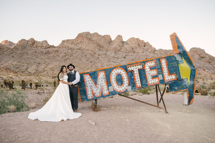 Bride in a satin ball gown and groom in a plaid vest and black hat stand beside the vintage MOTEL arrow sign at Nelson Ghost Town, rocky hills behind them.