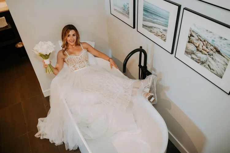 Bride in a lace sheath gown seated in a light-filled room holding a small white bouquet