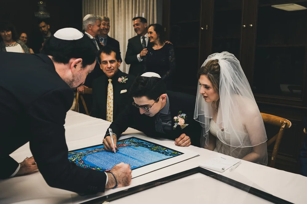 Groom signing the illuminated ketubah while the bride and the witnesses look on