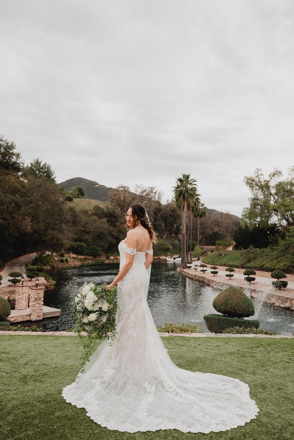 Bride in an off-the-shoulder lace fit-and-flare gown with a soft veil standing on a lawn beside a lake