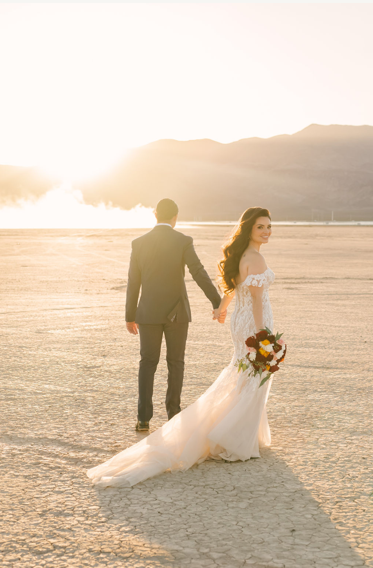 Bride in an off-the-shoulder lace gown looks back over her shoulder as she walks hand in hand with her groom across a cracked dry lake bed at sunset outside Las Vegas.