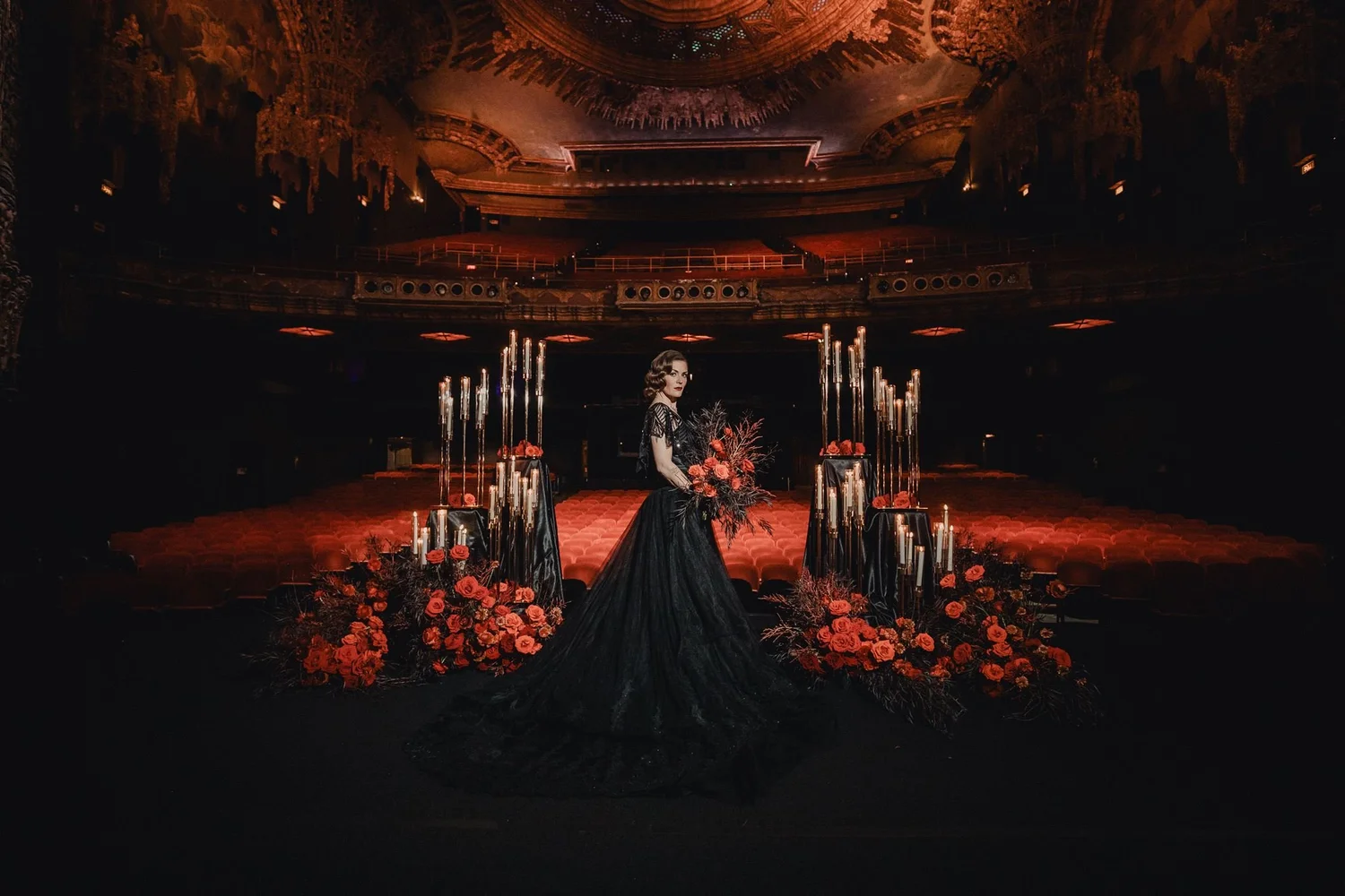 Bride in a black gown standing between tall arrangements of deep red roses and candles in a darkened historic theater