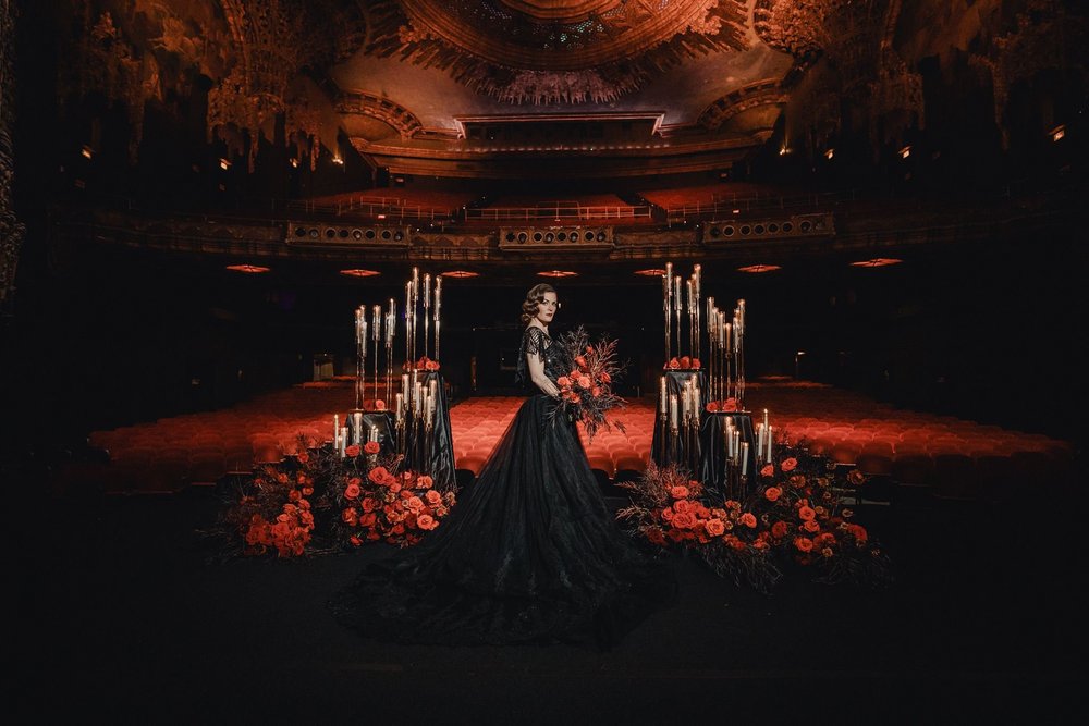 Wide portrait of a bride in a black wedding gown standing in a dark theatre space framed by tall arrangements of red roses