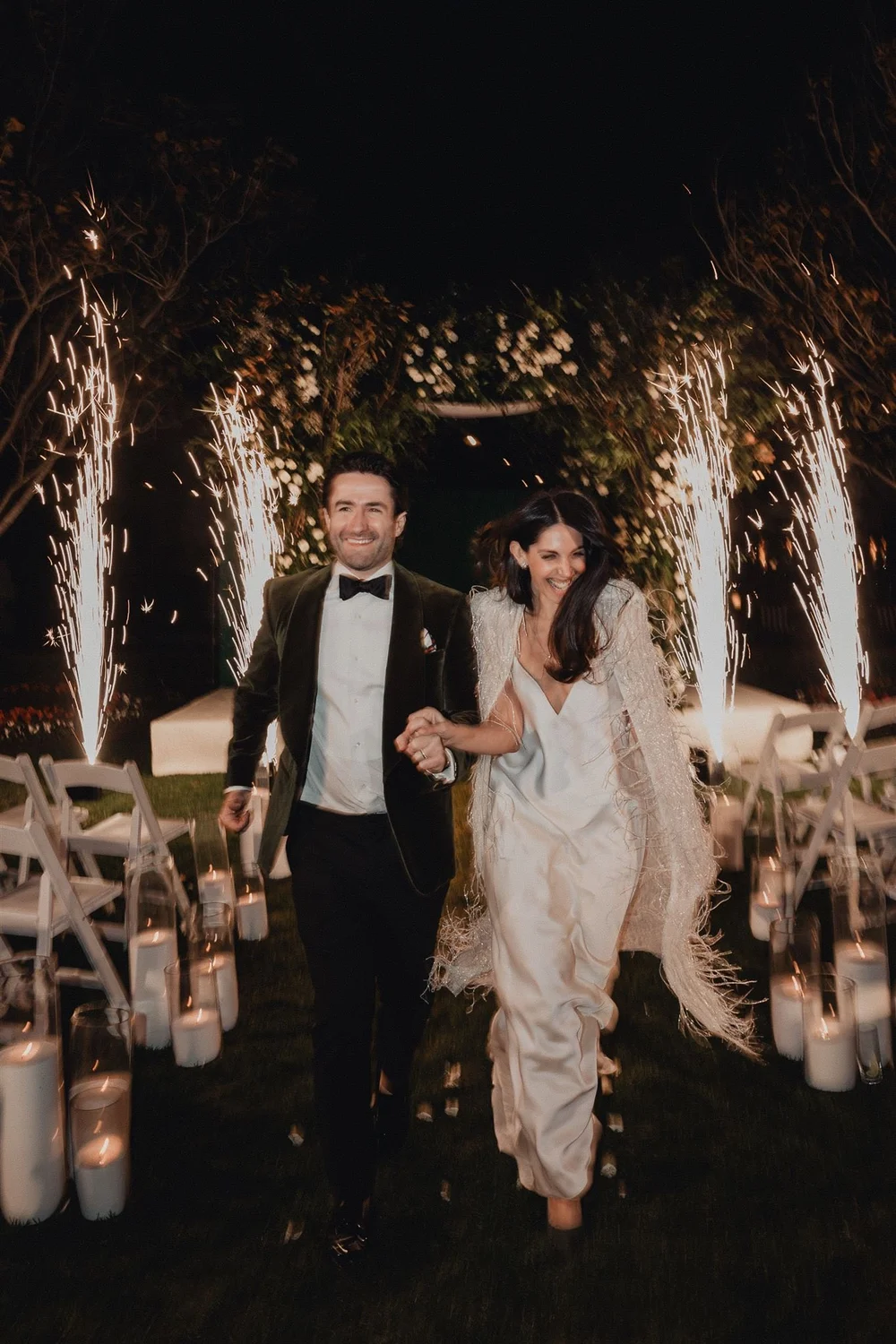 Couple walking out between cold-spark fountains at night, the candlelit ceremony aisle behind them at Rosewood Miramar Beach