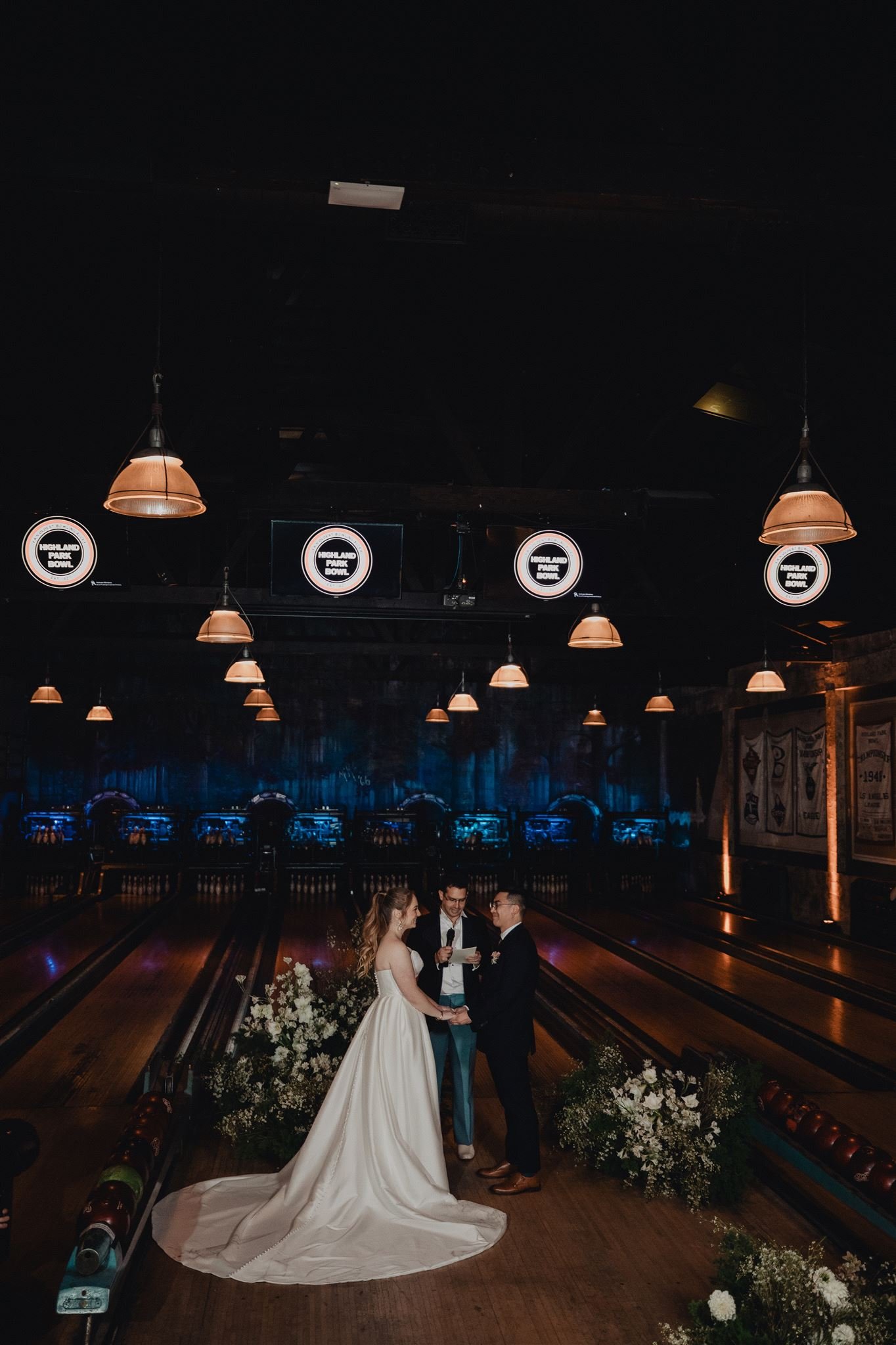 Wedding ceremony held on the lanes at Highland Park Bowl, floral arrangements either side and copper pendant lights overhead