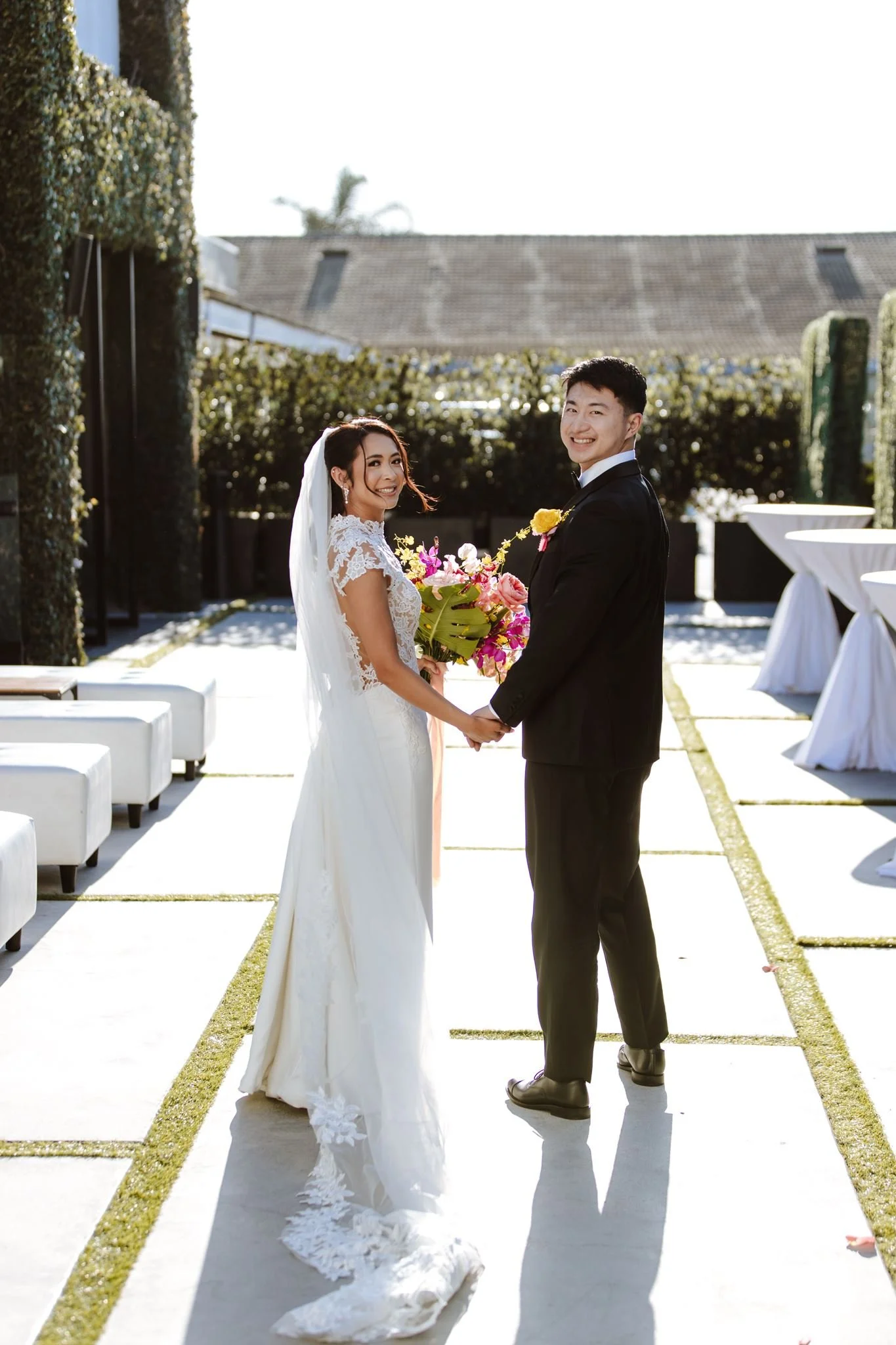 Couple holding hands on the sunny terrace with white lounge seating at Venue by Three Petals