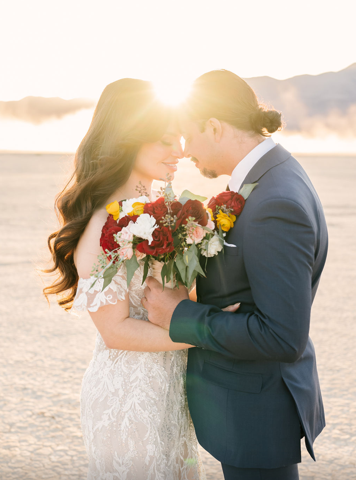 Bride and groom touch foreheads as the setting sun flares between them on a dry lake bed, a bouquet of red roses and yellow ranunculus held close.