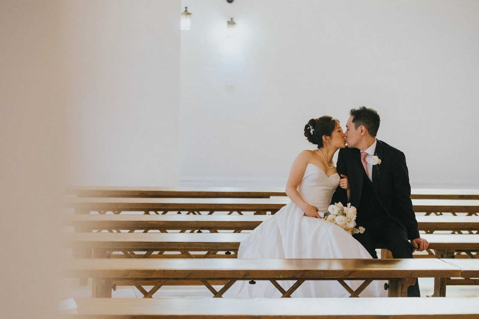 Couple kissing beside the light wood pews in the white chapel at The Sanctuary in Long Beach