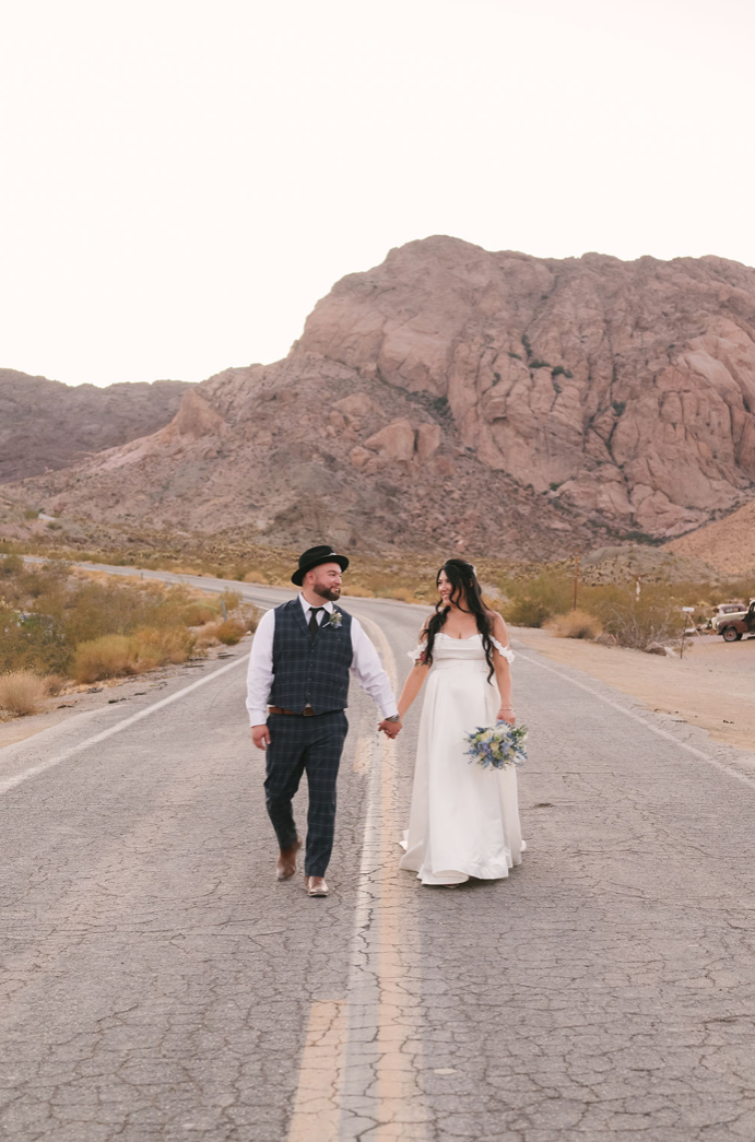 Bride and groom walk hand in hand down the center line of a cracked desert road below rugged rock peaks outside Nelson Ghost Town.