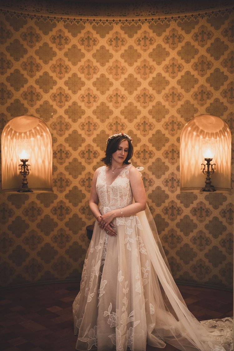 Bride in a champagne lace gown with a tulle overlay standing against patterned wallpaper between two wall lamps