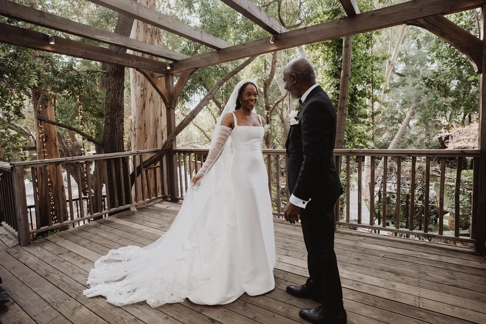 Bride in a lace-sleeved fit-and-flare gown with matching gloves turning to face her husband on a wooden deck