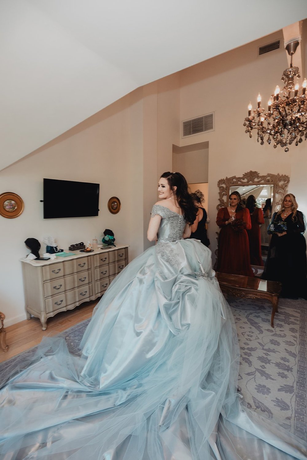 Bride in a pale blue tulle ball gown in a getting-ready suite with her bridesmaids arranging the skirt