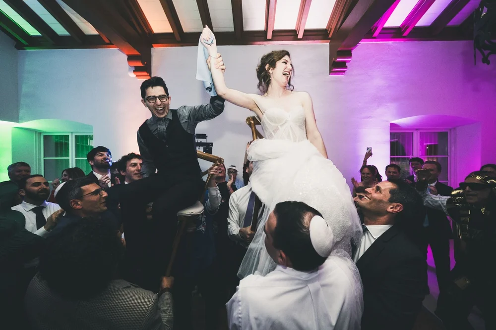 Bride and groom lifted on chairs holding a napkin between them under purple reception lighting
