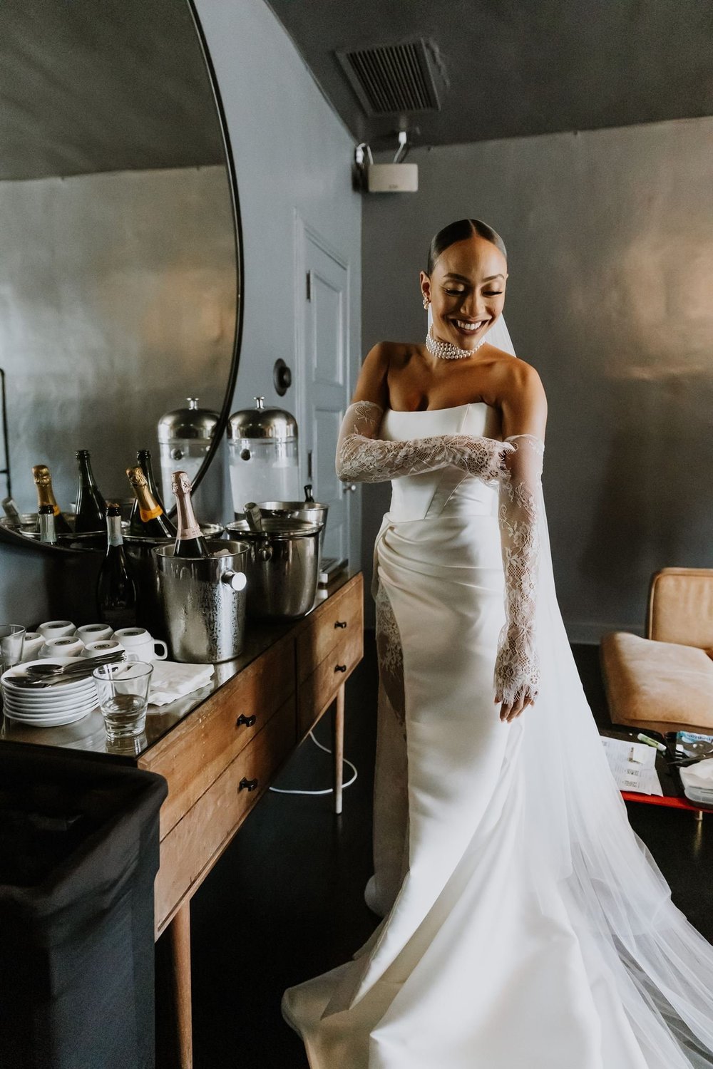 Bride in a fitted ivory gown wearing long textured gloves standing beside a bar in a dark panelled room