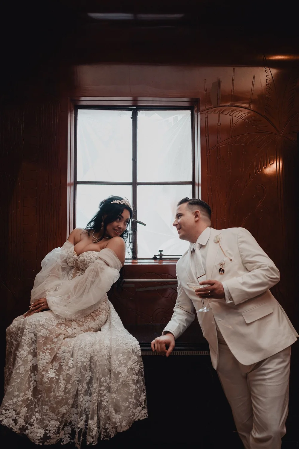 Couple in cream by the tall windows of the Oviatt Penthouse, the art deco woodwork behind them