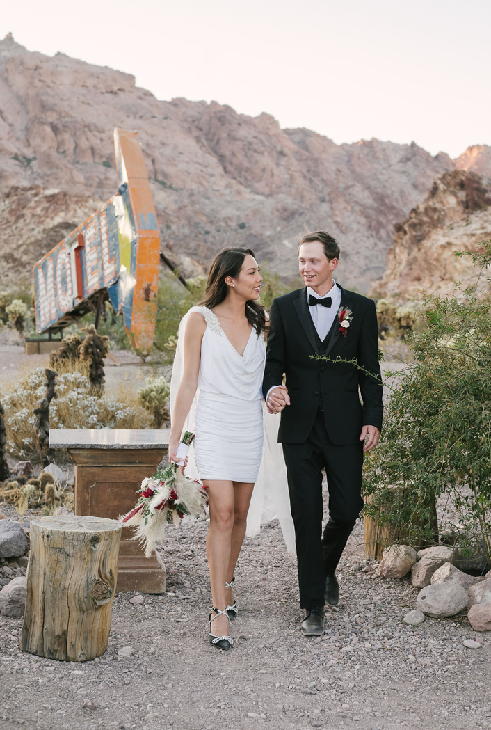Bride in a white mini dress and cape walks hand in hand with her groom in a tuxedo past a vintage sign at Nelson Ghost Town.