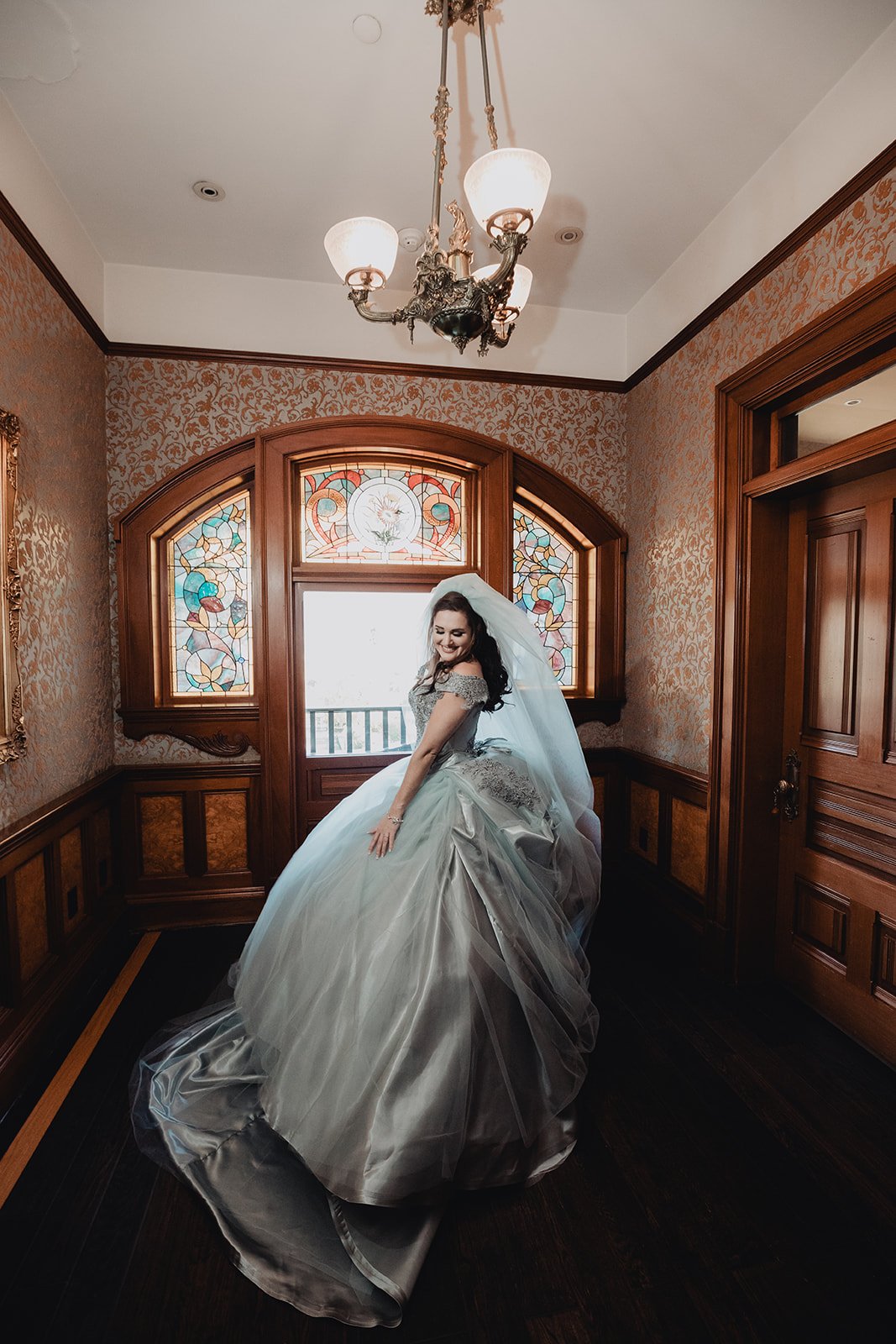 Bride sweeping a pale blue ball gown with a cathedral train through a wood-panelled hallway lit by stained glass