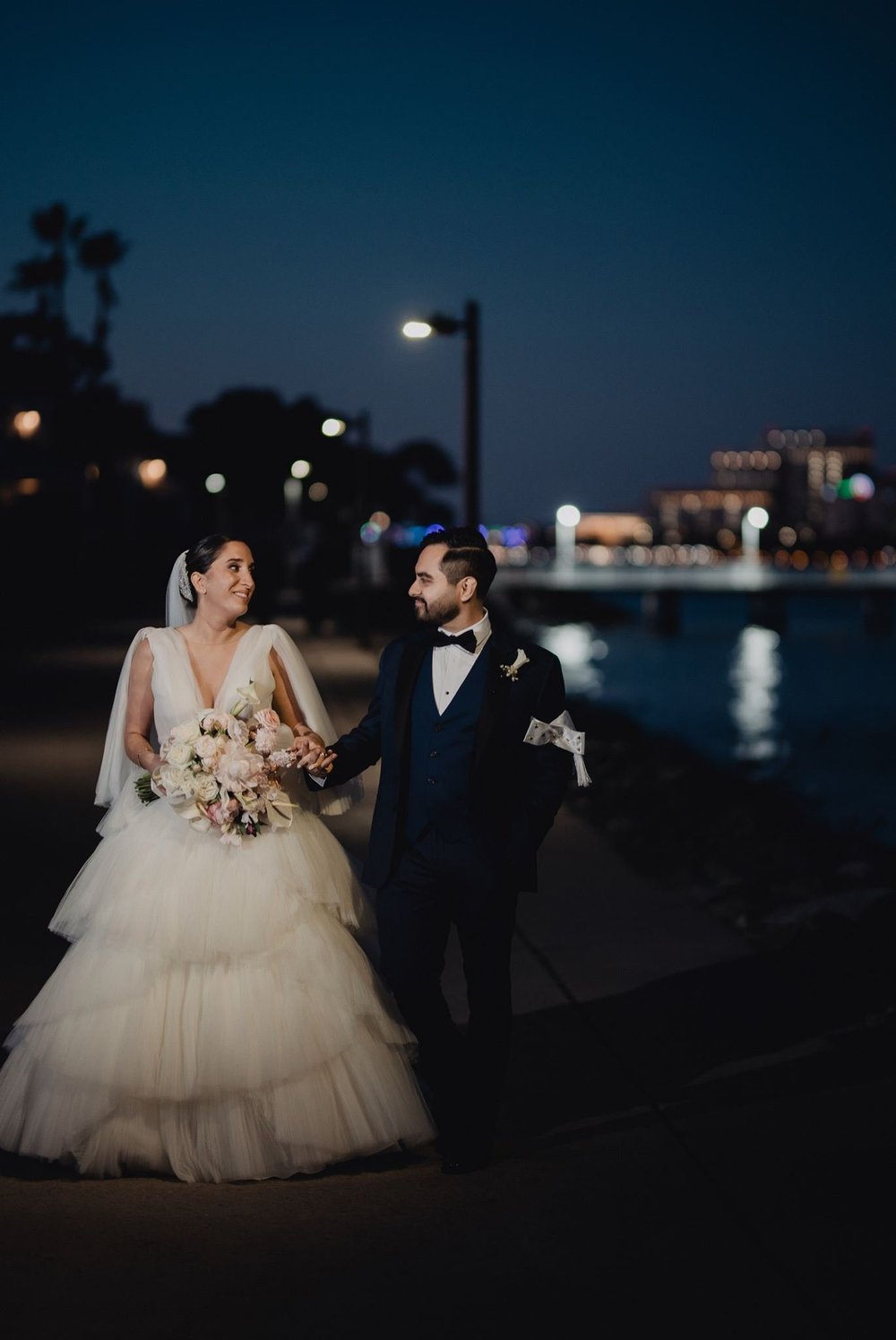 Bride in a tiered tulle gown with a corseted bodice walking with her husband beside a lake at night