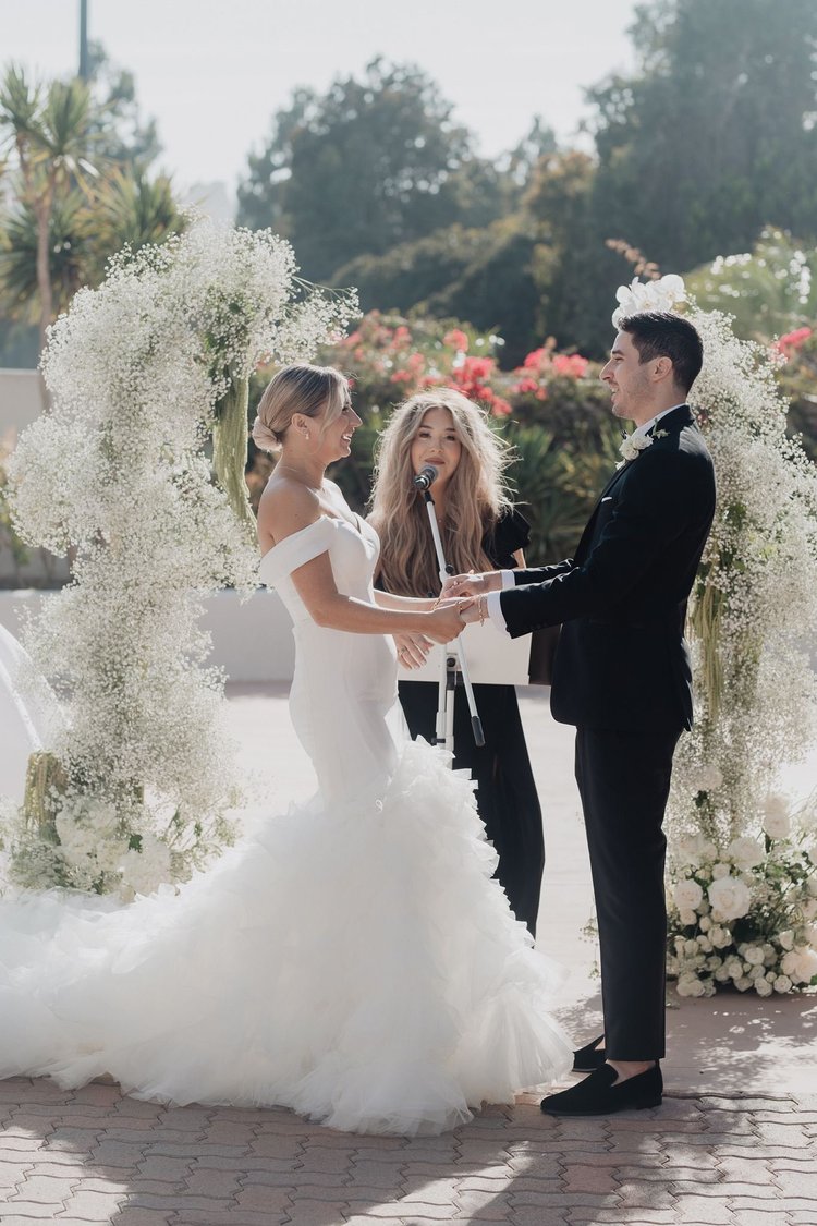 Bride in a heavily ruffled mermaid wedding dress at an outdoor ceremony beneath a floral arch
