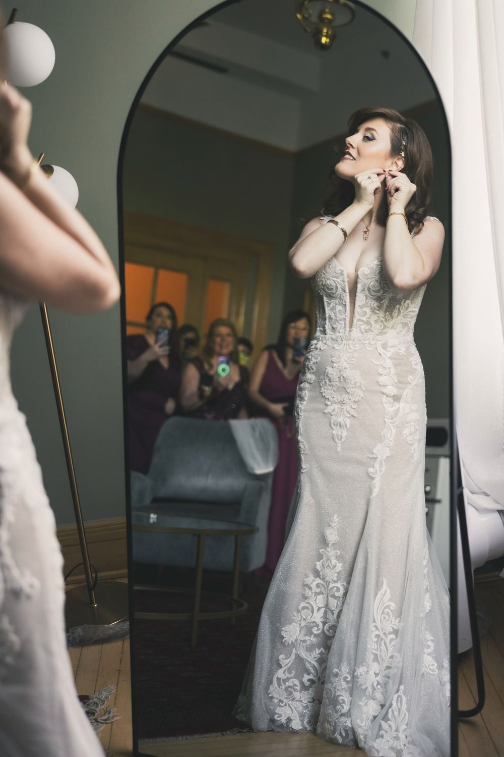 Bride in a fitted lace corset gown adjusting her hair in front of a large arched mirror
