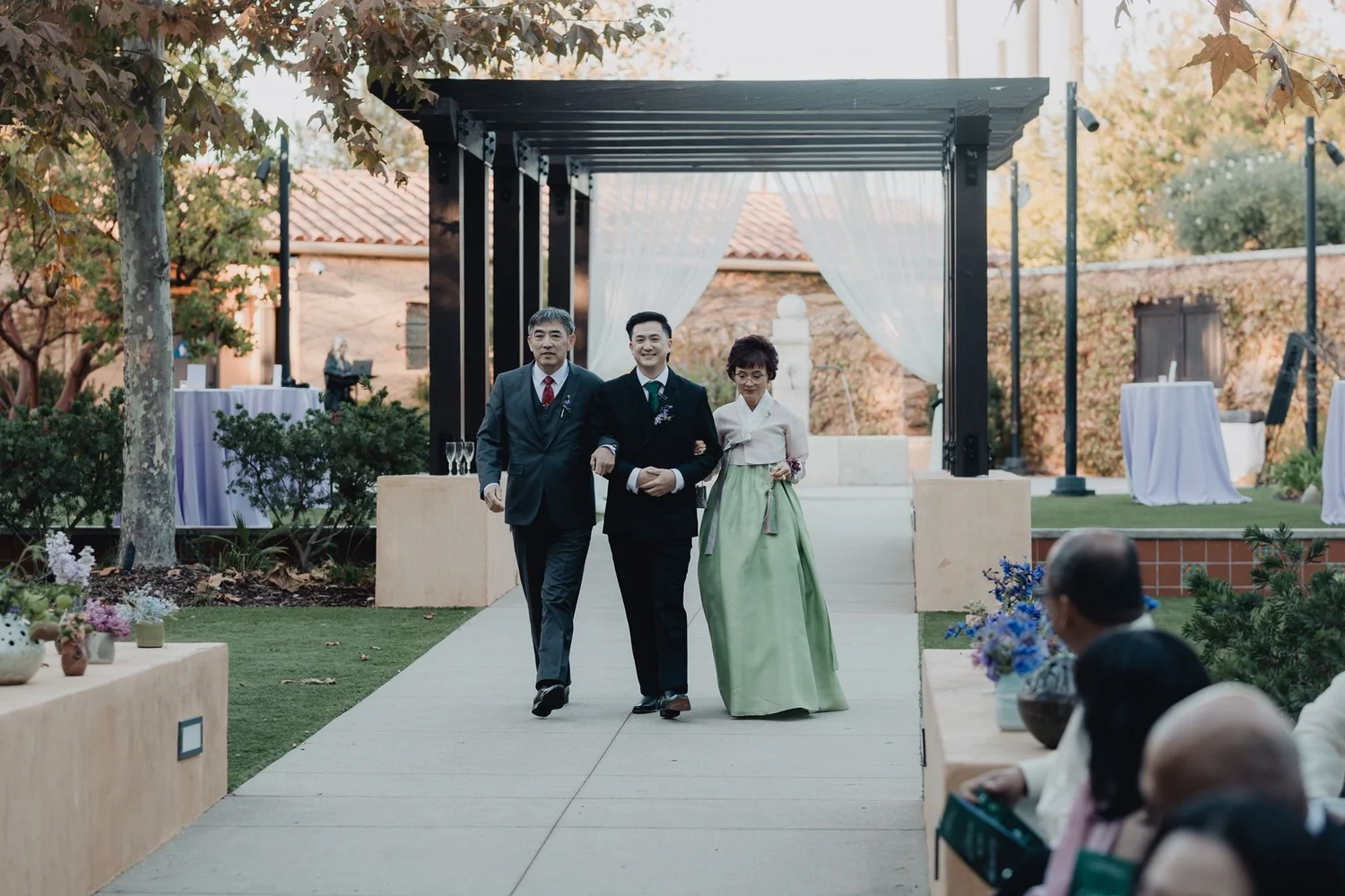 Korean family walking together under a garden pergola, one relative in a green hanbok, before the paebaek