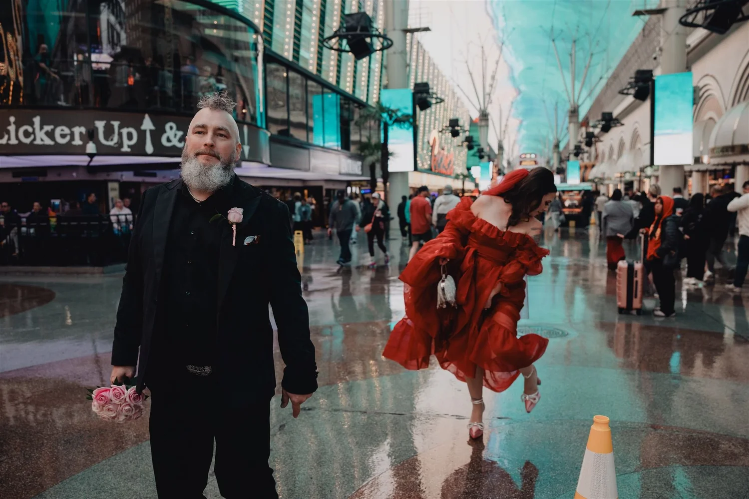 Groom in black holding a pink bouquet under the Fremont Street canopy while the bride in a red ruffled gown dances behind him, the Viva Vision screen glowing overhead
