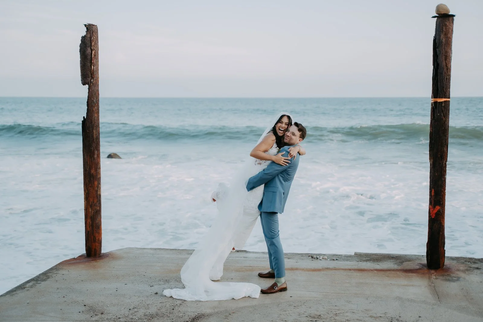 Groom lifting the bride on a concrete landing between old pilings above the surf at Duke's Malibu