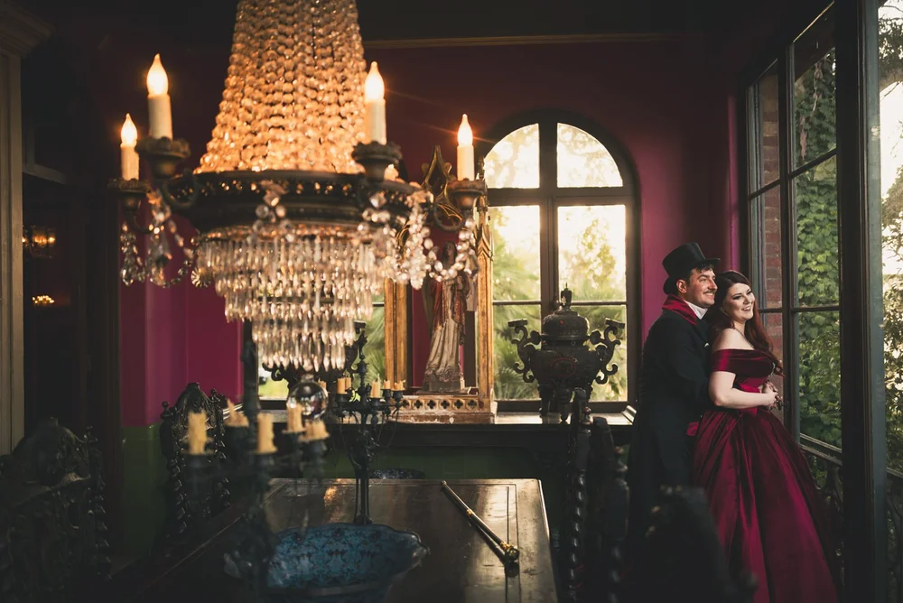 Couple in period dress beneath the crystal chandelier in a candlelit room at the Paramour Estate, the bride in a deep red gown