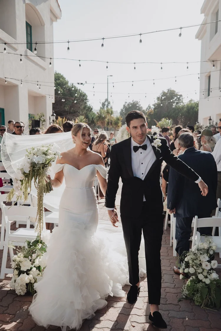 Bride in a heavily ruffled mermaid gown walking back up the aisle with her husband after the ceremony